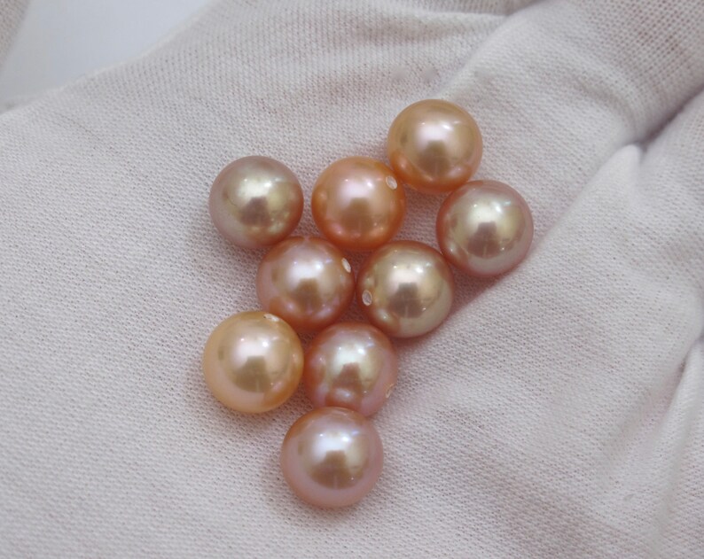 May include: A collection of lustrous, round pearls in varying shades of peach, pink, and gold. The pearls are displayed on a white fabric background, showcasing their smooth surfaces and natural iridescence. The pearls are of different sizes.