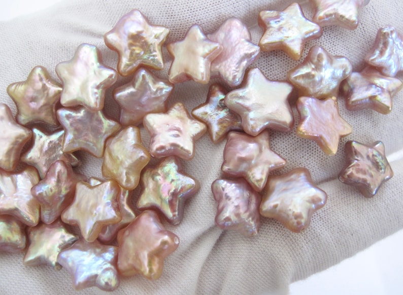 May include: A collection of iridescent pink star-shaped pearl beads. The beads are arranged in a loose pile on a white surface.