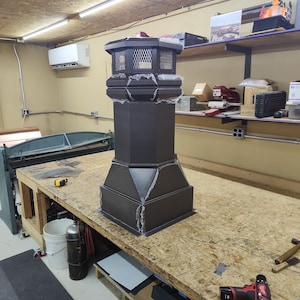 May include: A black metal chimney cap with a square base and a decorative top. The cap has a mesh screen on the top and is sitting on a wooden workbench.