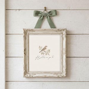 May include: A framed print with a bird illustration and the text "He watches over you too." The frame is ornate and off-white, with a sage green velvet bow above. The print is hung on a white wooden wall.