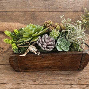May include: A rustic wooden planter box filled with a variety of faux succulents in shades of green, gray, and purple. A small animal skull is attached to the arrangement. The box has a dark brown finish and metal handles.