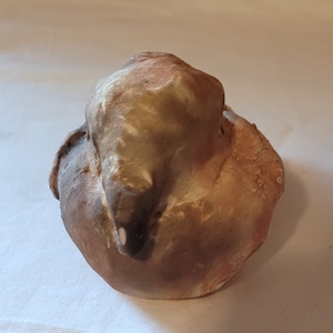 Puede incluir: Una pequeña escultura de pájaro de cerámica con un esmalte marrón, blanco y gris. El pájaro está sentado con la cabeza gacha y las alas plegadas.