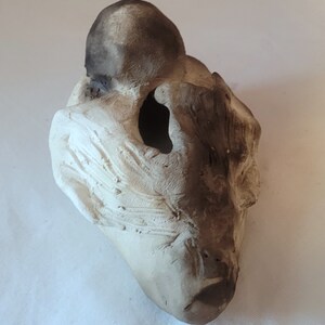 May include: A white ceramic sculpture of a face with a dark gray top. The sculpture has a hole in the center of the face and is textured.