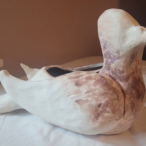 May include: A white ceramic bird planter with brown and purple painted accents. The bird is lying on its side with its wings spread out. The planter has a small opening on the top for planting.
