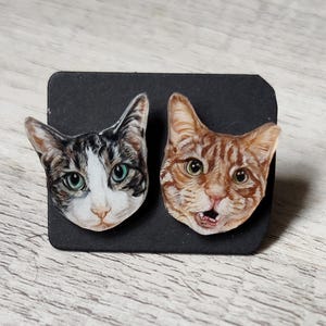 May include: Two cat face stickers. One cat has gray and white fur with green eyes. The other cat has orange and brown fur with an open mouth. Both stickers are on a black background.