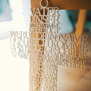May include: A wooden cross with the Lord's Prayer carved into it. The cross is made of light-colored wood and has a circular top. The text is in German and is cut out of the wood.