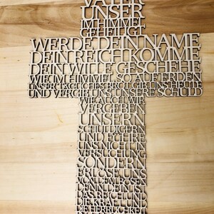 May include: A wooden cross with the Lord's Prayer in German laser-cut into the wood. The cross is light brown and has a small hole at the top for hanging. The text is in a sans-serif font.