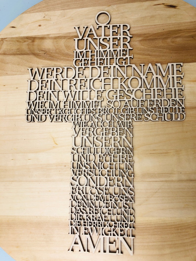 May include: A wooden cross with the Lord's Prayer in German laser-cut into the wood. The cross is light brown and has a small hole at the top for hanging. The text is in a sans-serif font.