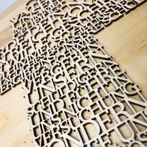 May include: Wooden cross with cut-out letters. The cross is made of light-colored wood, with the letters forming the shape of the cross. The letters are in a sans-serif font and appear to be German. The background is a light wood grain.