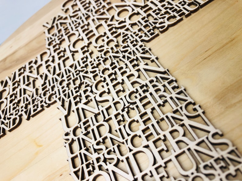 May include: Wooden cross with cut-out letters. The cross is made of light-colored wood, with the letters forming the shape of the cross. The letters are in a sans-serif font and appear to be German. The background is a light wood grain.
