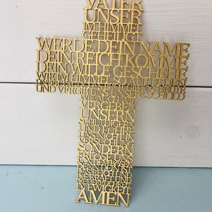 May include: A wooden cross with the Lord's Prayer inscribed in German. The cross is gold-colored and has a loop at the top for hanging. The text is cut out of the wood, creating a unique design.