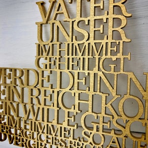May include: A gold-colored wooden cross with the German text of the Lord's Prayer cut out. The cross has a loop at the top for hanging. The background is a white wooden surface.