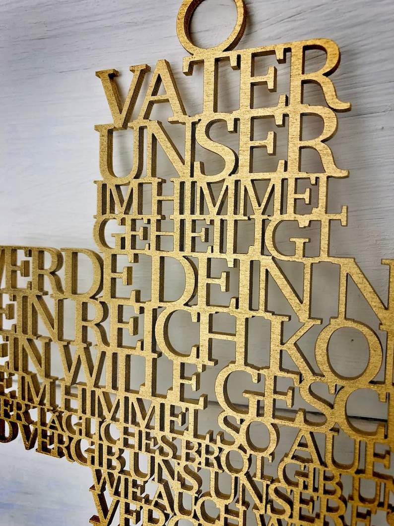 May include: A gold-colored wooden cross with the German text of the Lord's Prayer cut out. The cross has a loop at the top for hanging. The background is a white wooden surface.