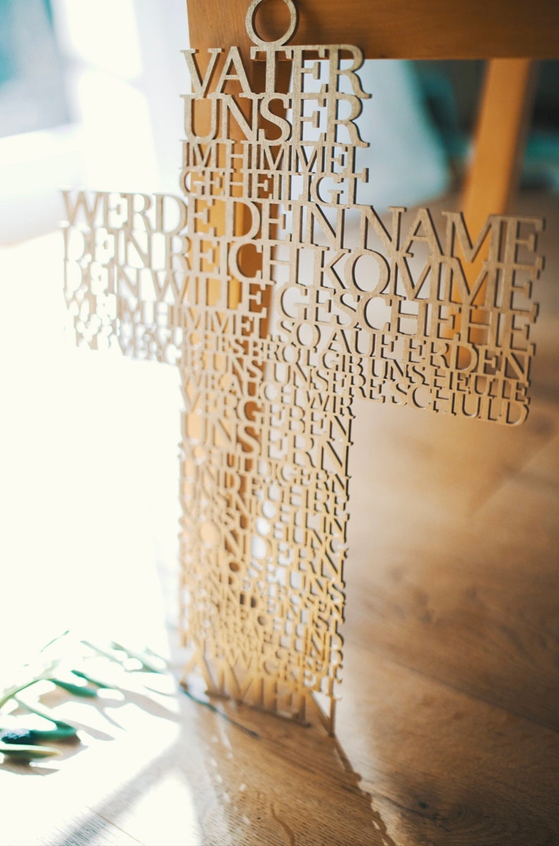 May include: A wooden cross with the Lord's Prayer carved into it. The cross is made of light-colored wood and has a circular top. The text is in German and is cut out of the wood.