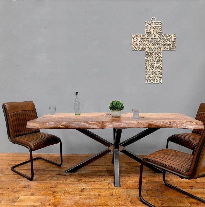 May include: A wooden dining table with a live edge top and a black metal base. Two brown leather chairs with black metal frames are on either side of the table. A wooden cross with German text hangs on the gray wall.