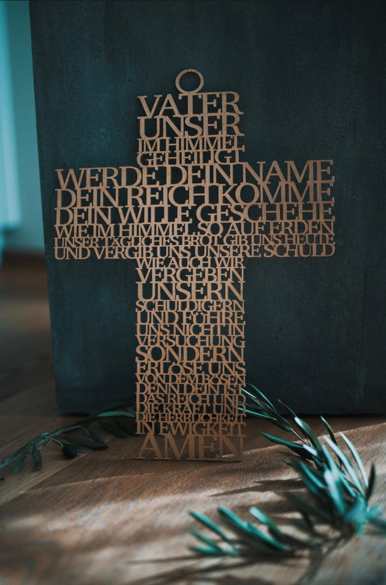 May include: A wooden cross with the Lord's Prayer etched into it. The cross is made of light brown wood and is set against a dark gray background. The text is in German. The word "Amen" is at the bottom.