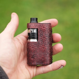 May include: A brown leather vape mod with a floral pattern. The mod is being held in a person's hand.