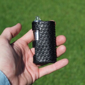 Puede incluir: Un mod de vape de cuero negro con un patr&oacute;n texturizado. El mod se sostiene en una mano.