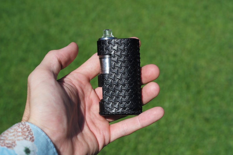 Puede incluir: Un mod de vape de cuero negro con un patr&oacute;n texturizado. El mod se sostiene en una mano.