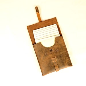May include: A brown leather notepad holder with a flap closure and a strap. The holder has a white notepad inside and the text "Gentlemen's Goods" embossed on the front.