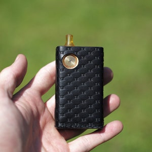 Op de afbeelding: Een zwarte leren vape-mod met een gouden knop en een doorzichtige drip tip.