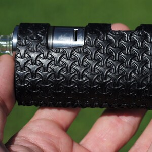 Puede incluir: Un mod de vape de cuero negro con un patr&oacute;n geom&eacute;trico. El mod tiene una parte superior de metal plateado y una punta de goteo de vidrio transparente.