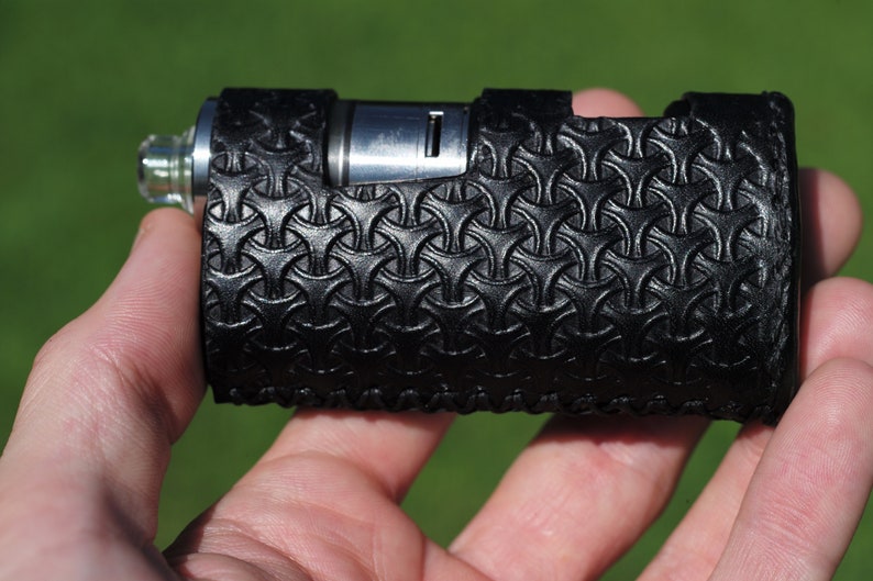 Puede incluir: Un mod de vape de cuero negro con un patr&oacute;n geom&eacute;trico. El mod tiene una parte superior de metal plateado y una punta de goteo de vidrio transparente.