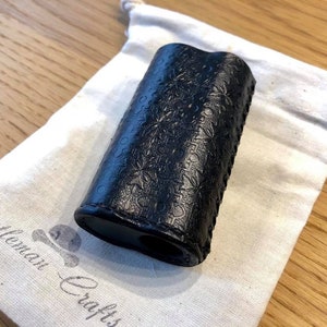 May include: A black leather lighter case with a floral embossed design. The case is sitting on a white fabric bag with the text "Gentleman Crafts" printed on it.