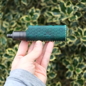 May include: A black vape pen with a green leather sleeve. The sleeve has a geometric pattern and the words "Gentleman's Crafts" stitched on it. The vape pen is held in a hand against a blurred green background.