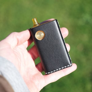 May include: A black leather vape mod with a gold button and a clear drip tip. The mod is stitched with white thread around the edges.