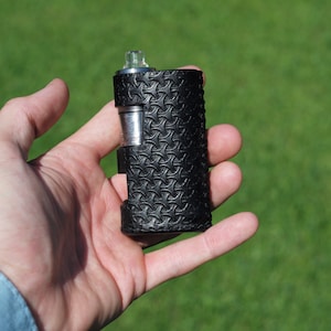 Puede incluir: Un mod de vape de cuero negro con un patr&oacute;n texturizado. El mod tiene una parte superior plateada y un tanque de vidrio transparente.