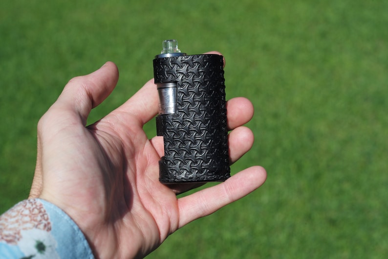 Puede incluir: Un mod de vape de cuero negro con un patr&oacute;n texturizado. El mod tiene una parte superior plateada y un tanque de vidrio transparente.