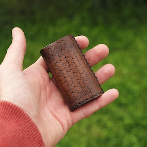 May include: A brown leather case with a geometric pattern, held in a hand. The case has a stitched edge and is made of a textured leather.