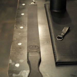 Puede incluir: Una correa de afeitar de cuero negro con un mango cosido y un logotipo en relieve en la parte delantera. La correa está sobre una superficie metálica.