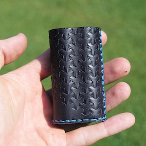 May include: A black leather case with a geometric pattern and blue stitching. The case is held in a hand.