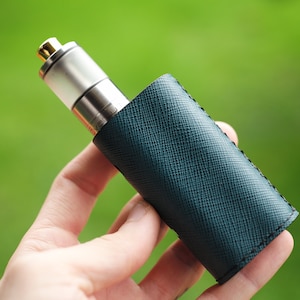 May include: A silver and gold vape pen is partially encased in a dark teal leather sleeve. The sleeve has a textured surface and is stitched along the edges. The background is a blurred green.