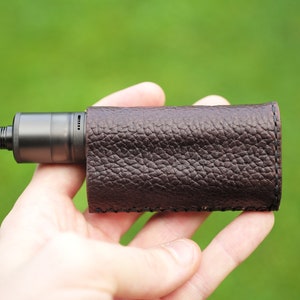 May include: A brown leather case for an electronic cigarette. The case is made of a textured leather and has a stitched seam around the edges.