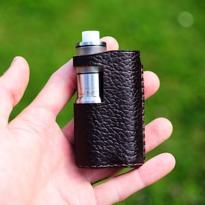 May include: A brown leather case with a silver vape mod inside. The case has a textured leather finish and a stitched seam around the edges.