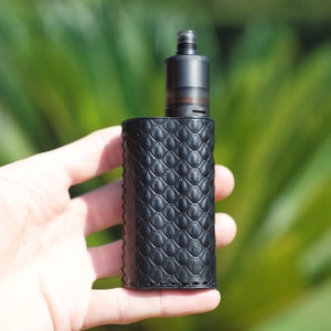 May include: A black vape mod with a textured, snake-skin-like design. The mod is held in a hand.