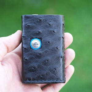 Può includere: Un portafoglio in pelle nera con un motivo in pelle di struzzo. Il portafoglio ha un piccolo bottone metallico con un anello blu intorno.