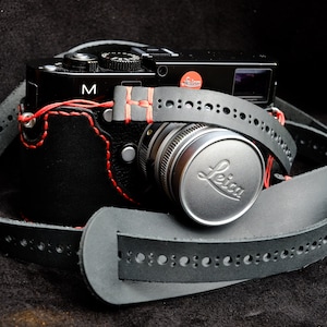 Puede incluir: Una correa de cámara de cuero negro con costuras rojas y una tapa de lente plateada con la palabra "Leica" grabada. La correa de cámara tiene un patrón decorativo de pequeños agujeros.