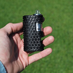 Puede incluir: Un mod de vape de cuero negro con un atomizador plateado y una punta de goteo. El mod tiene un patr&oacute;n texturizado.