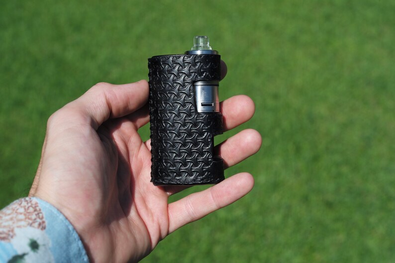 Puede incluir: Un mod de vape de cuero negro con un atomizador plateado y una punta de goteo. El mod tiene un patr&oacute;n texturizado.