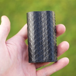 May include: A black leather case with a scale-like pattern. The cylindrical case is held in a hand against a blurred green background. The case appears to be made of high-quality leather and is stitched along the edges.