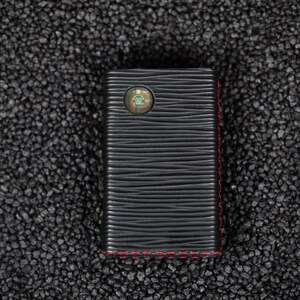May include: A black leather case with red stitching and a small circular cutout on the front.