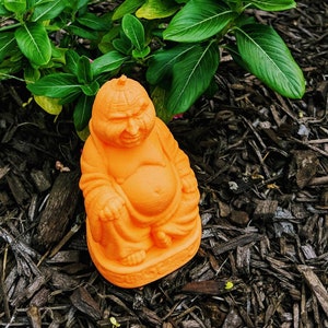 Puede incluir: Estatua de cerámica naranja de un Buda sonriente sentado con las manos en el regazo. La estatua está sobre un lecho de virutas de madera marrón.