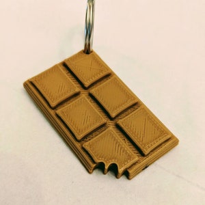 May include: A brown chocolate bar keychain with a silver key ring. The chocolate bar is broken off at one end.