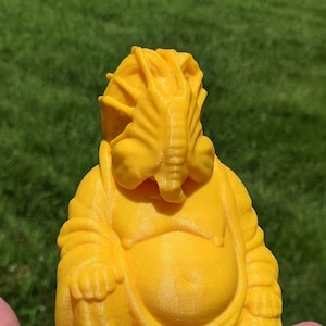 Puede incluir: Una figura de Buda sonriente de color amarillo impresa en 3D con una cabeza de extraterrestre. El Buda está sentado en una postura tradicional con las manos en el regazo. La cabeza extraterrestre tiene un diseño detallado con crestas y una boca.