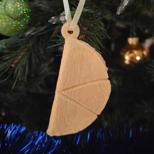 Puede incluir: Un adorno navideño de color beige, impreso en 3D con forma de porción de pizza con tres rebanadas. El adorno está colgado de una cinta blanca en un árbol de Navidad verde.