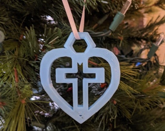 May include: A light blue heart-shaped Christmas ornament with a white cross inside. The ornament is hanging on a Christmas tree.